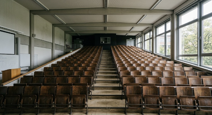 Leerer Hörsaal von vorn gesehen: ansteigende Reihen hölzerner Klappsitze beiderseits eines Mittelgangs, rechts eine durchgehende Fensterfront, links Tafel und Pult